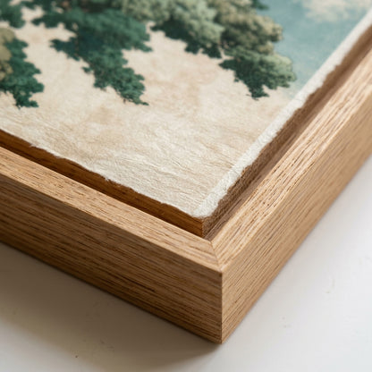Close-up of a framed art print showing the corner of a landscape painting with green trees on textured off-white paper, set in a light oak wood frame.