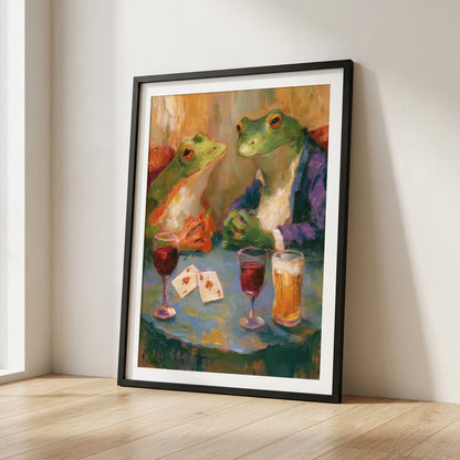 A framed art print leaning against a wall on a light wood floor. The print shows two green frogs seated at a table with drinks and playing cards, rendered in a painterly, colorful style.